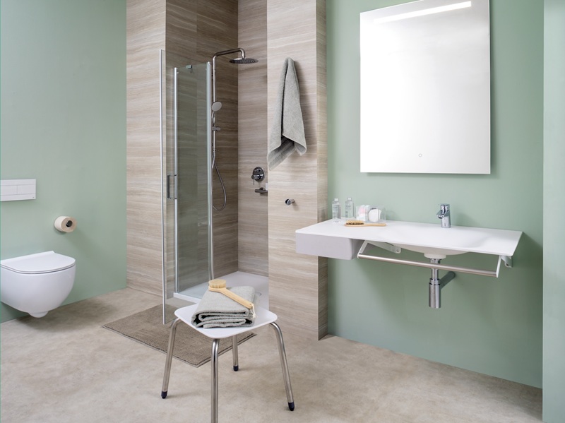 Bagno accessibile con doccia in box di vetro, lavabo sospeso con maniglione, sgabello e pareti verde salvia
